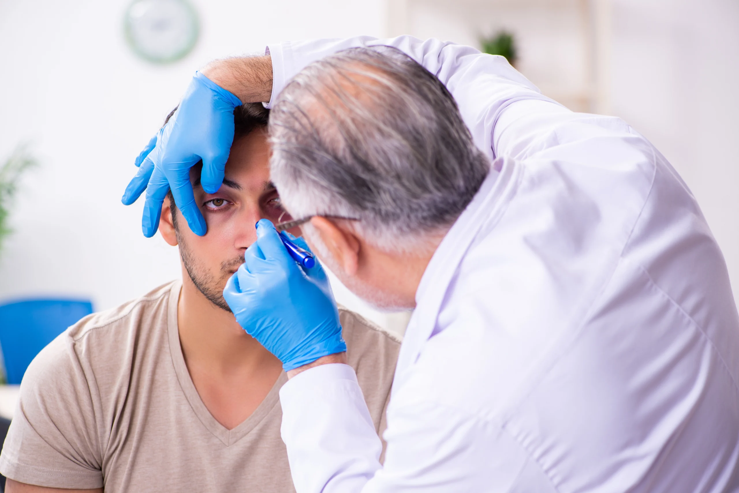 A young man receives an eye exam from an older doctor wearing a lab coat and blue gloves.